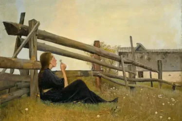 una ragazza siede in un prato, appoggiata a una staccionata, mentre sta per soffiare un soffione di un tarassaco.