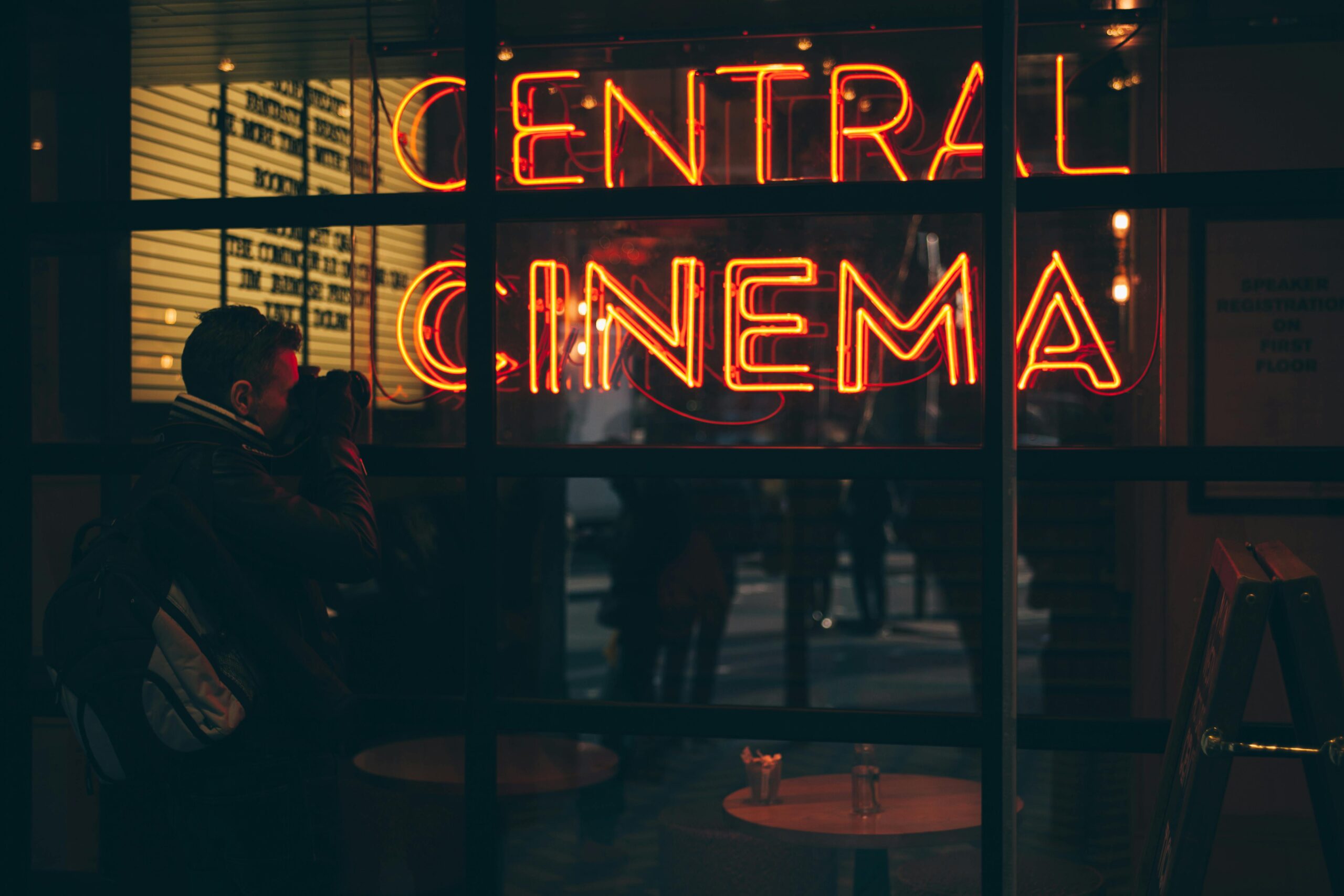 un uomo scatta una foto ad una vetrina dove si riflette la scritta in neon arancione "Central Cinema".